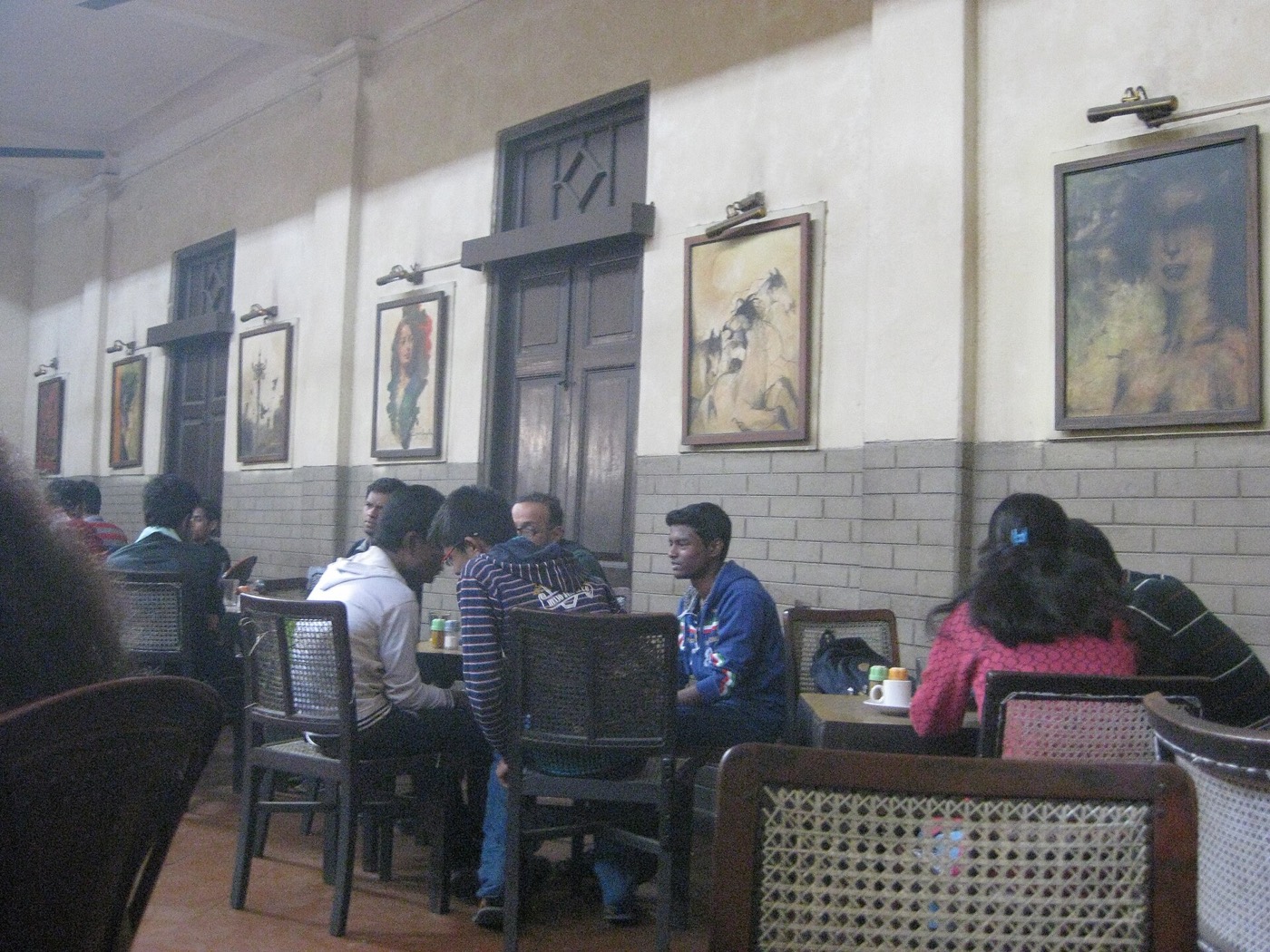 Salão amplo de um café antigo em Calcutá, com pé direito alto, retratos nas paredes e várias mesas ocupadas por grupos conversando